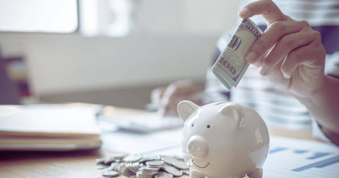 A hand placing a hundred dollar bill in a piggy bank.